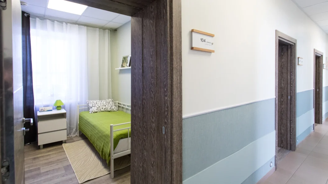 Corridor with bedrooms. There is an open door with a bed with green sheets and a matching lamp on a white bedside table.