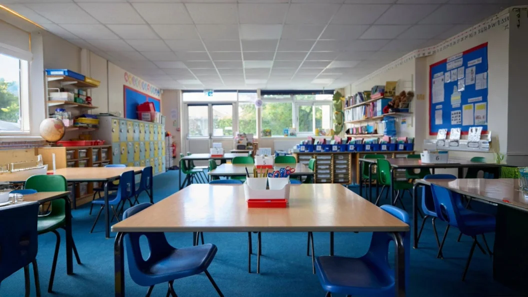 Interiors of a primary school classroom in England with tables and blue and green chairs.